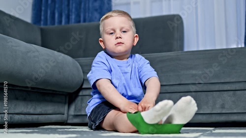 A young child is sitting on the floor of a living room. The child is holding green tape, looking focused while playing. The couch and curtains are visible in the background.