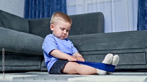 A child sits on the floor and pulls a resistance band with both hands while sitting on a rug in a living room. Sunlight shines through a nearby window.