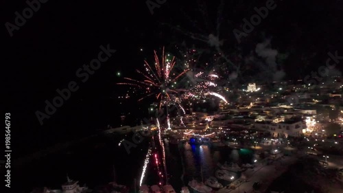 A cinematic FPV drone flight through colorful fireworks over a coastal town at night. Bright explosions illuminate the sky creating a festive and dynamic atmosphere