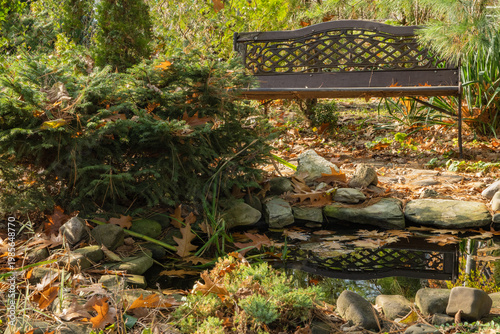 Decorative brown bench sits above small, rocky pond surrounded by lush evergreens, fallen leaves, and tall grasses, reflecting tranquility in garden.