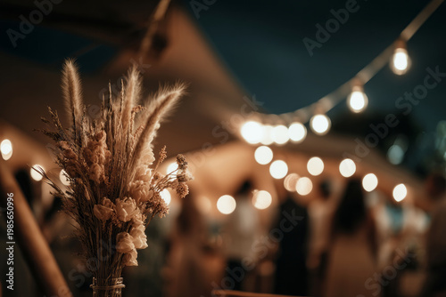 Guests gather under a large wedding tent filled with warm lights. The area is decorated with flowers and creates a festive ambiance for the outdoor celebration at dusk