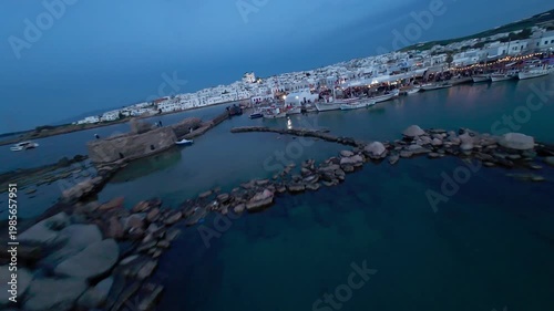 Cinematic FPV drone flight over Naousa village on Paros island during sunset, featuring warm evening light, coastal harbor and Easter celebrations with festive atmosphere