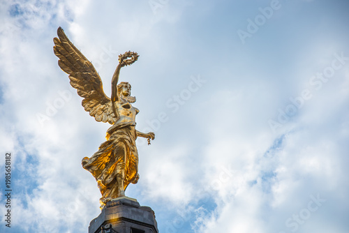 Low-angle view of the gilded Angel of Independence atop its column on Paseo de la Reforma in Mexico City, holding a laurel wreath and broken chains against a partly cloudy sky.