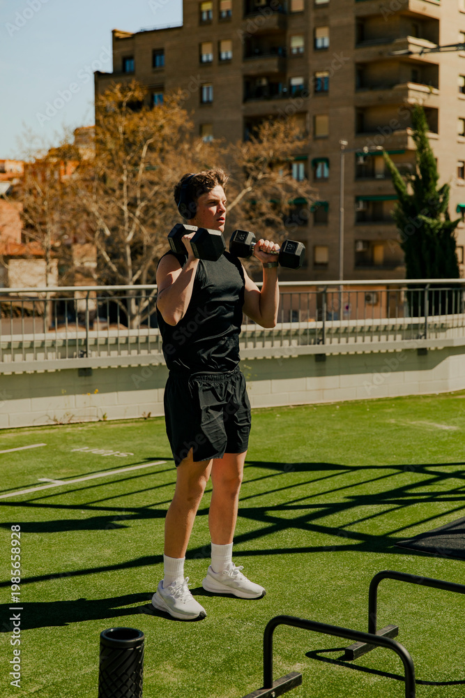 Fototapeta premium Fit young man lifting dumbbells overhead during strength workout in outdoor gym. Fitness training, power and endurance concept.
