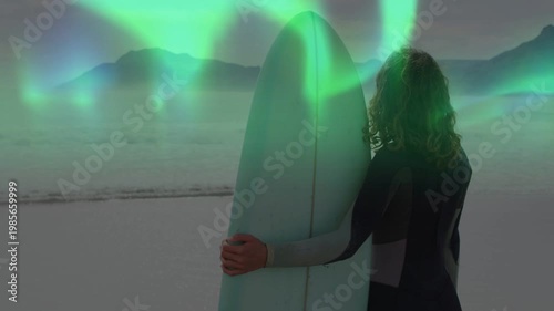 Vertical video: Teal aurora sweeping causing surfer holding board turning toward lights for ads