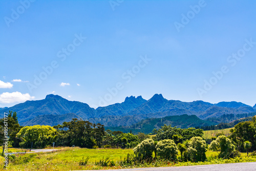 Sunny rural landscape with green fields and winding road leading toward rugged mountain peaks under a vast blue sky, capturing freedom, distance, and natural beauty. Pauinui, New Zealand
