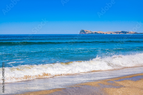 A vibrant turquoise wave curls and breaks onto a sandy shore under a clear blue sky. Refreshing energy and beauty of a pristine coastal seascape. Pauinui, Coromandel Peninsula, New Zealand