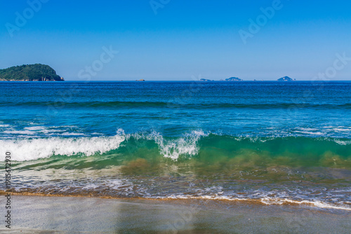 A vibrant turquoise wave curls and breaks onto a sandy shore under a clear blue sky. Refreshing energy and beauty of a pristine coastal seascape. Pauinui, Coromandel Peninsula, New Zealand