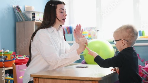 Young boy wearing glasses participates in educational activity with teacher, colorful learning cards spread on the table in a bright classroom environment