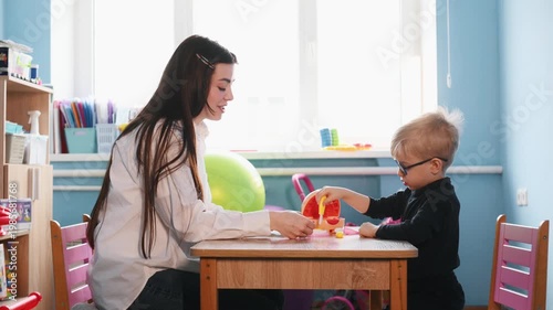 Preschool child interacts with teacher during playtime at a wooden table, surrounded by colorful toys and classroom elements in a bright indoor environment