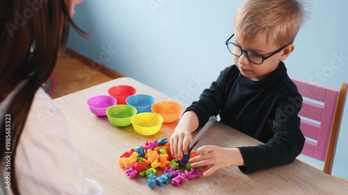 Young child wearing glasses engages in cutting colorful fruit pieces on a table, while a teacher is visible in the background assisting with the activity