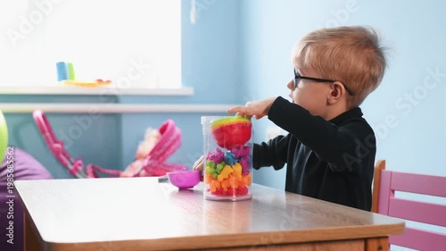 Preschool boy wearing glasses focuses on placing colorful shapes into a clear container on a wooden table, surrounded by bowls of various colors