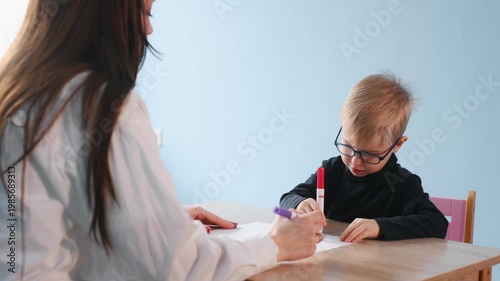 Preschool child works on a drawing with a teacher at a wooden table in a bright classroom, featuring educational toys and colorful decor