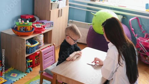 Young boy wearing glasses participates in educational activity with teacher, colorful learning cards spread on the table in a bright classroom environment