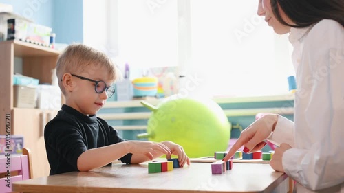Young boy wearing glasses plays with colorful building blocks on a wooden table, while a teacher's hand assists him in a bright classroom environment