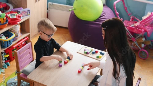 Young boy with glasses interacts with colorful blocks on a table while a teacher guides him in a bright classroom environment