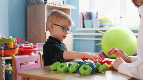 Young boy wearing glasses plays with colorful building blocks on a wooden table, while a teacher's hand assists him in a bright classroom environment