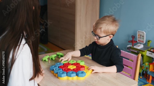 Preschool boy with glasses plays with colorful building blocks on a table while a teacher assists him in a vibrant classroom filled with educational toys