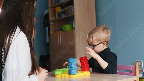 Young boy wearing glasses arranges colorful building blocks on a table while a teacher guides him in a well-lit classroom environment