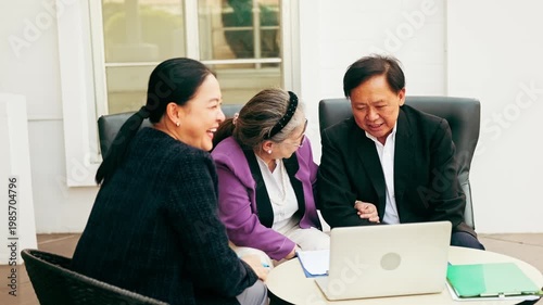 Three adults sit together around a laptop, smiling and discussing paperwork during an informal business meeting.
