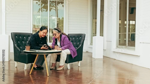 Two women sit on outdoor lounge chairs by a white building, sharing a tablet and coffee while discussing work.