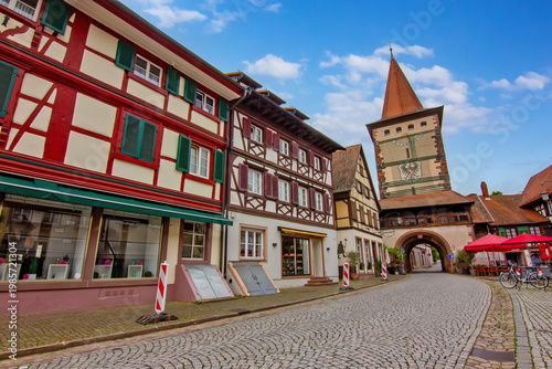 Colorful historic half timbered houses along cobbled street in Gengenbach old town with tower gate and flowers under blue sky