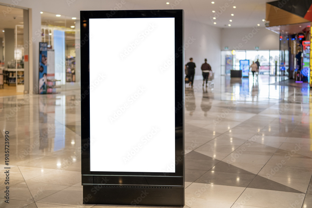 Obraz premium A blank white vertical digital billboard lightbox display stand mockup in a modern Australian shopping centre mall corridor, with blurred shoppers and retail stores in the background. Template for ads