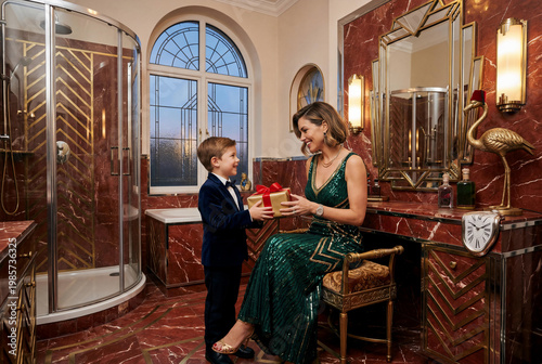 Young boy in a velvet suit giving a gift to his elegant mother in a green sequin gown within a luxury red marble bathroom featuring art deco mirrors and surrealist decor for Mother's Day.