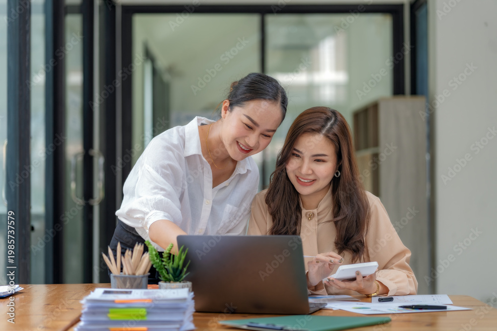 Fototapeta premium Two Asian businesswomen collaborating and discussing financial project on laptop and documents in office.