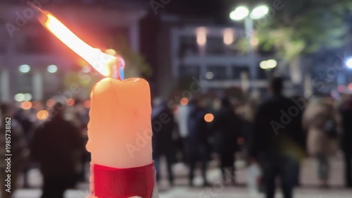 Burning candle in sharp focus with blurred crowd holding lights in the background during an Easter nighttime outdoor gathering.