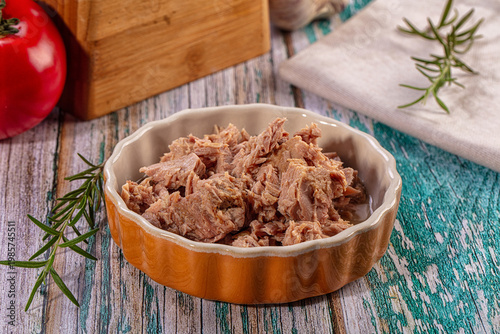 Flaked canned tuna in an orange ceramic bowl with fresh rosemary and tomato on a rustic wooden table