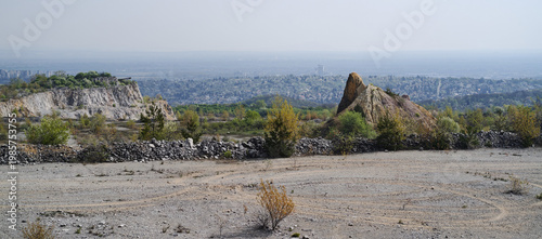 Wide view of an abandoned quarry with unique rock formations and sparse vegetation. Miskolc city stretches in the background under soft hazy light.