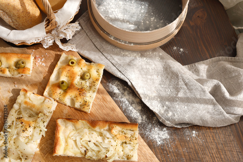 Artisan Italian focaccia slices with olives and onions on wooden board with traditional flour sieve. High-angle shot features variety of homemade breads pink Himalayan salt and wicker basket under