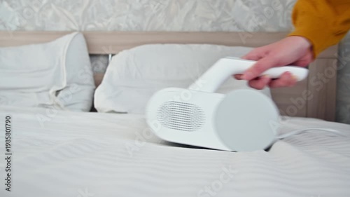 Handheld vacuum cleaner removes dust from bed linens in bright bedroom setting