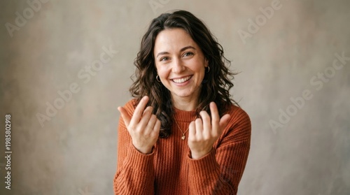 Warmly smiling brunette woman in rust orange sweater beckons with her hands, inviting gesture against a textured grey studio background, ideal for friendly marketing campaigns.