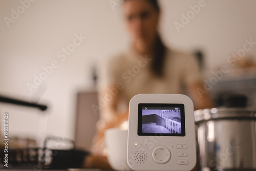 Digital baby monitor showing a sleeping infant in a crib on a kitchen counter, blurred young mother cooking in the background, multitasking parent concept, modern childcare and domestic lifestyle