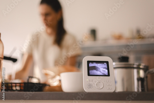 Digital video baby monitor with infant on screen placed on a kitchen counter, blurred young mother multitasking in the background, modern parenting technology for home safety and child surveillance
