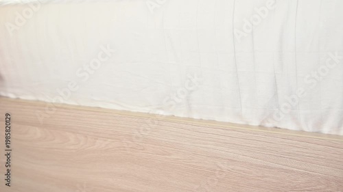 Hand tucks white bed sheet under wooden bed frame in bright bedroom setting