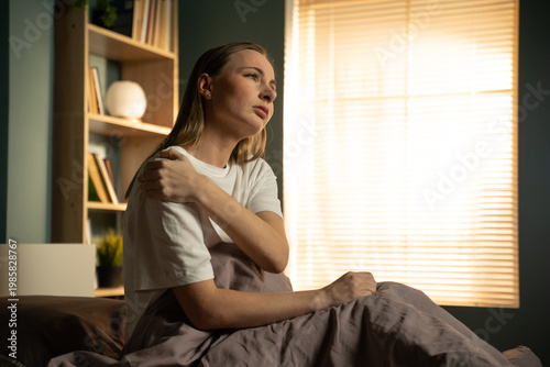 Woman sitting in bed, holding her shoulder in discomfort, feeling an injury or muscle ache after waking up, highlighting concepts of pain, exhaustion, and healthcare