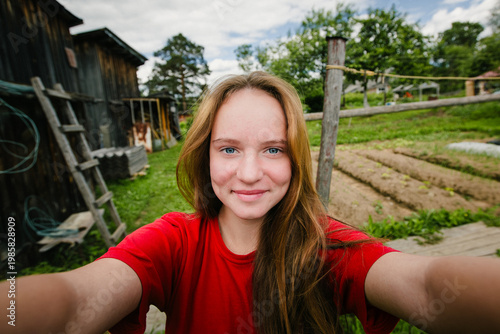 Young woman taking a selfie in a rural garden with wooden house and vegetable beds in the background.