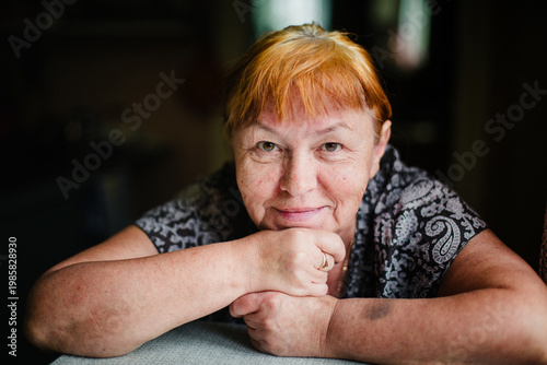 Middle aged woman resting her chin on hands, looking at camera, soft natural light, intimate indoor portrait.