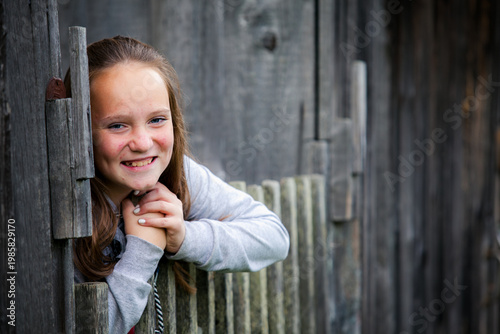 Smiling girl leaning on a wooden fence by a rustic house, outdoor portrait with soft natural light.