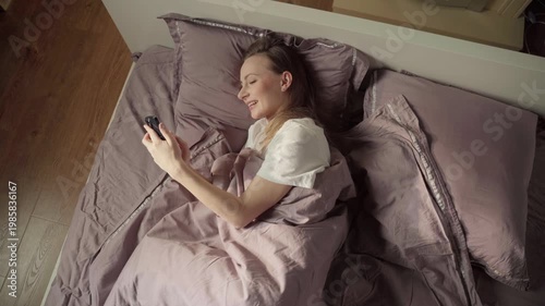 High angle view of a smiling young woman relaxing in bed and browsing her social media feed on a mobile phone after waking up in the morning, enjoying a peaceful start to her day