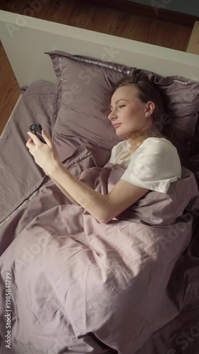 Happy young woman lying in a comfortable bed under a warm blanket in the morning, smiling as she scrolls through social media on her smartphone and checking messages before getting up