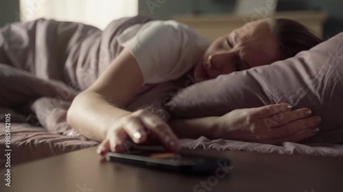 Annoyed sleepy woman turning off her smartphone alarm in the early morning, wanting to continue sleeping in her cozy bed instead of waking up, showing the difficulty of early rising