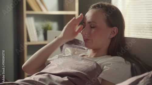 Unhealthy young woman suffering from a painful headache, high fever, and migraine symptoms while lying in bed, feeling sick and unwell with flu or a cold in her sunlit bedroom