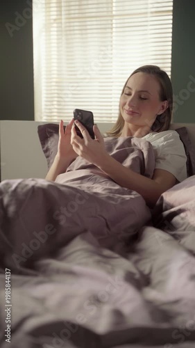 Charming young woman sitting in her bed in the morning, smiling while scrolling through her social media feed on a smartphone and enjoying a relaxed start to the day in her cozy bedroom