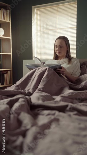 Peaceful young woman relaxing in her comfortable bed in the morning, enjoying a captivating book and smiling while turning the pages in a cozy, sunlit bedroom during a leisurely weekend
