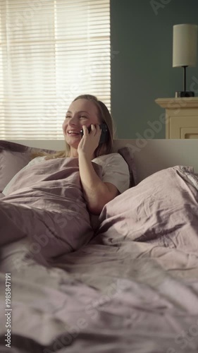 Happy young woman laughing and gesturing while having a pleasant conversation on her mobile phone, comfortably lying in bed in a cozy, sunlit bedroom during the morning