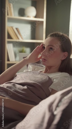 Stressed young woman lying in bed at home, suffering from a severe headache, migraine, or fever, gently massaging her temples to relieve the pain and discomfort of her illness